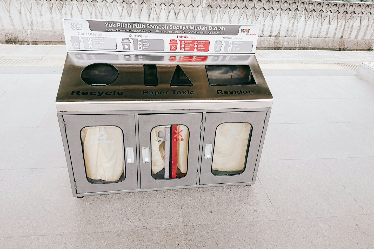 Ein Metall-Recyclingbehälter mit beschrifteten Fächern steht auf dem Boden.